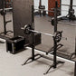 Black power squat stand with adjustable barbell safety arms and weight plate storage, shown in gym setting with loaded barbel