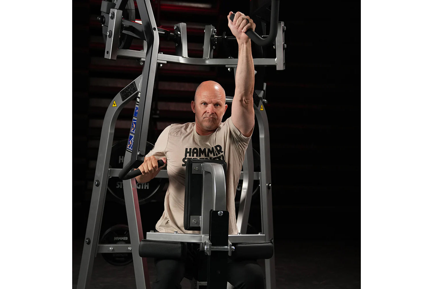 Man using gray and black Hammer Strength iso-lateral high row machine, wearing beige t-shirt with "HAMMER" logo, demonstratin