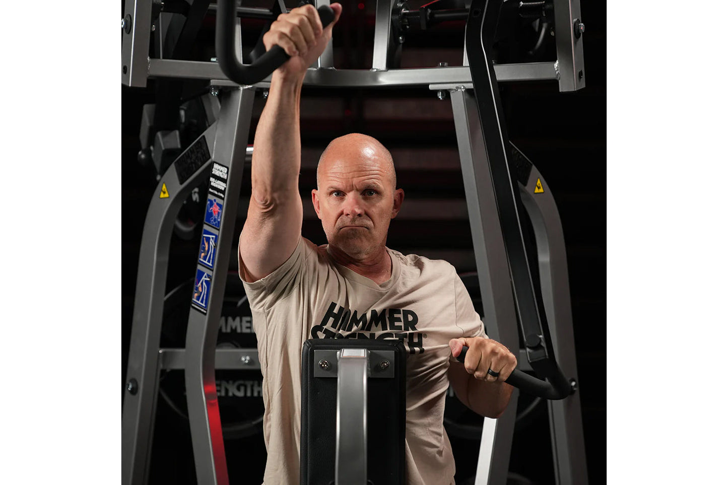 Man wearing beige Hammer Strength t-shirt using black Hammer Strength iso-lateral high row machine with arms extended overhea