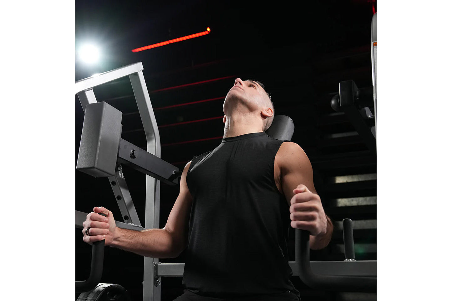 Man using black Hammer Strength plate-loaded neck exercise machine in gym setting with red neon lighting