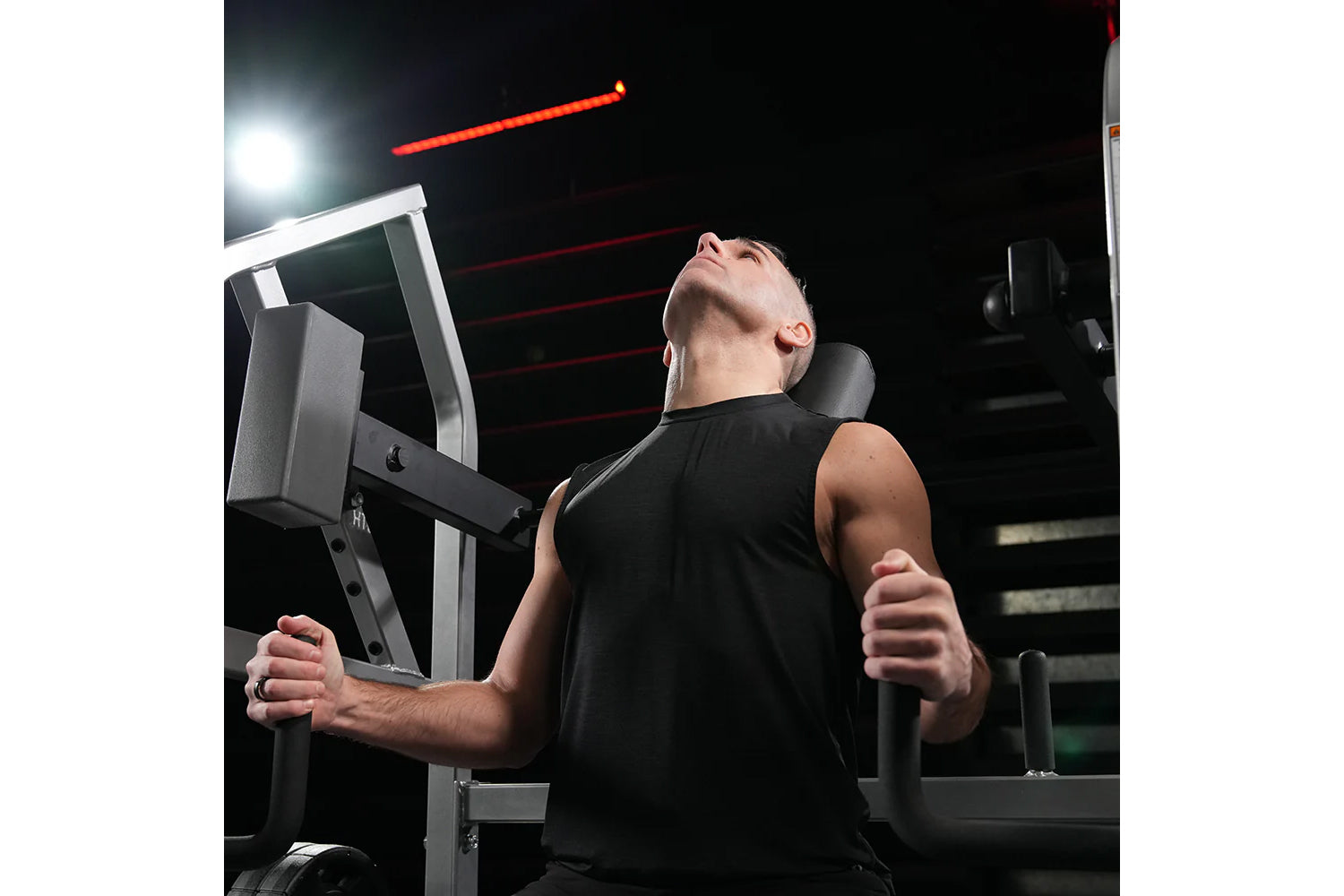 Man using black Hammer Strength plate-loaded neck exercise machine in gym setting with red neon lighting
