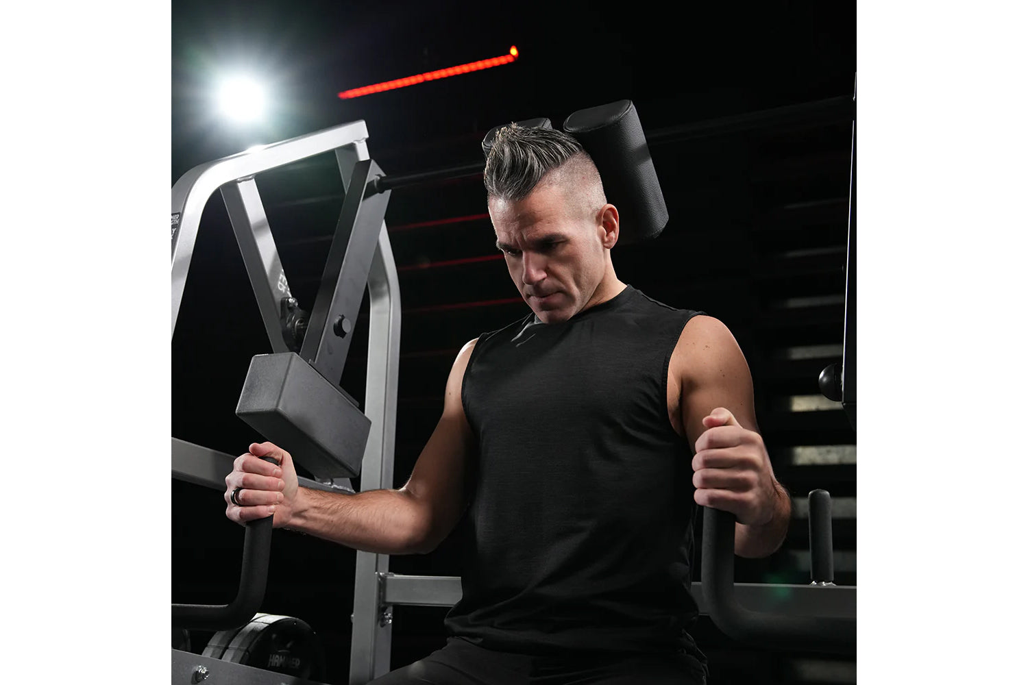 Man wearing black sleeveless tank top using gray Hammer Strength plate-loaded neck exercise machine in gym setting