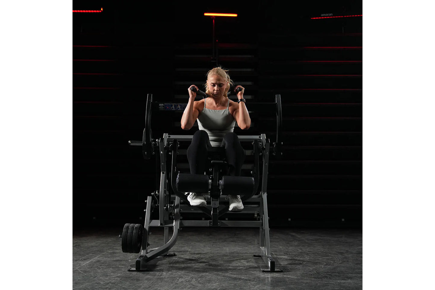 Woman performing abdominal crunch on Hammer Strength plate-loaded machine, wearing gray tank top and black leggings in gym se