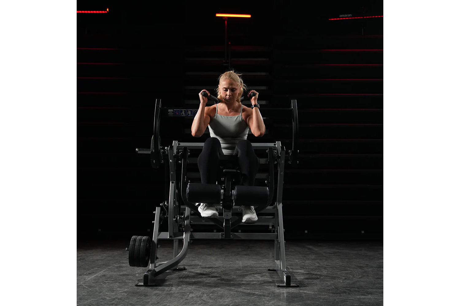 Woman performing abdominal crunch on Hammer Strength plate-loaded machine, wearing gray tank top and black leggings in gym se