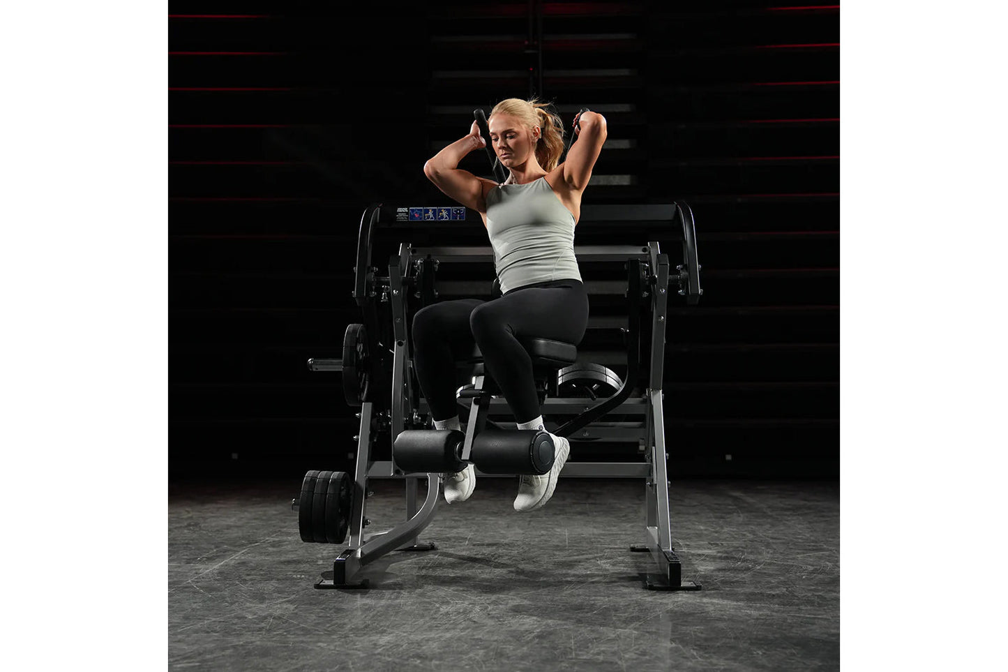 Woman using Hammer Strength plate-loaded abdominal oblique crunch machine in gym, wearing light gray tank top and black leggi