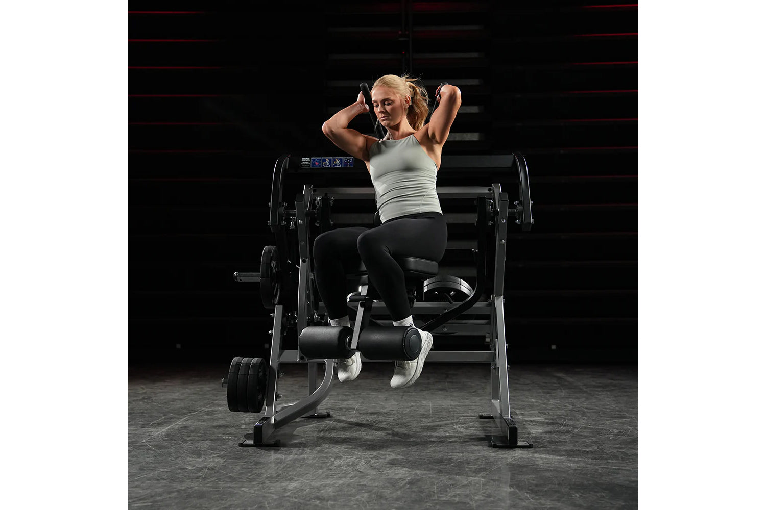 Woman using Hammer Strength plate-loaded abdominal oblique crunch machine in gym, wearing light gray tank top and black leggi