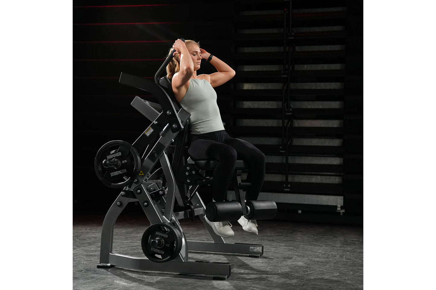 Woman using black Hammer Strength plate-loaded abdominal oblique crunch machine in gym setting, wearing gray tank top and bla