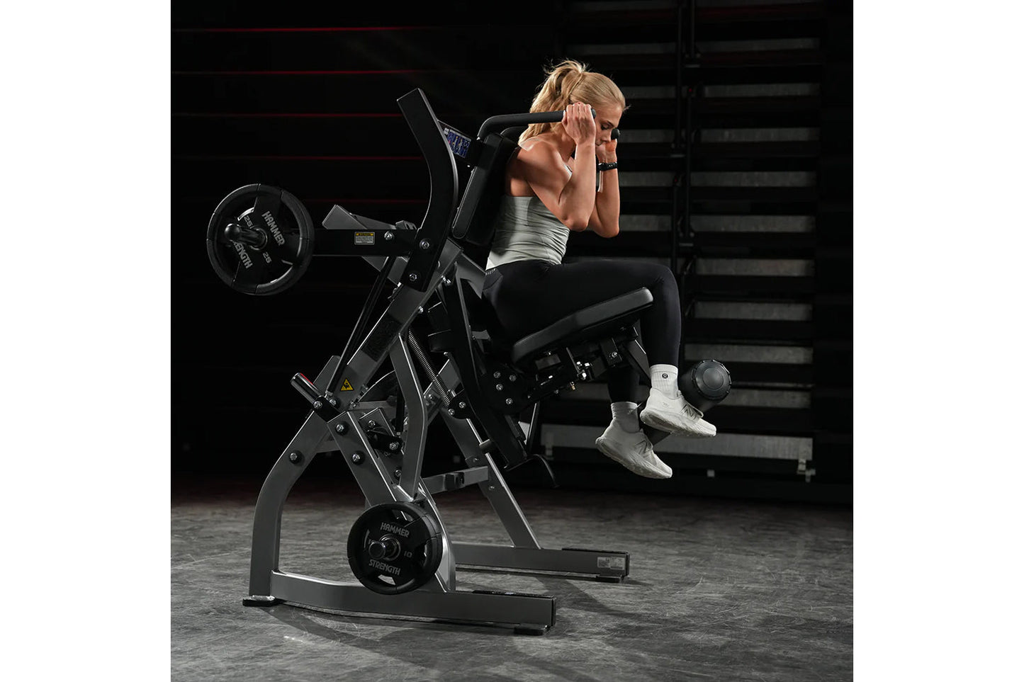 Woman using black plate-loaded abdominal oblique crunch machine in gym setting, wearing silver sports top and black leggings