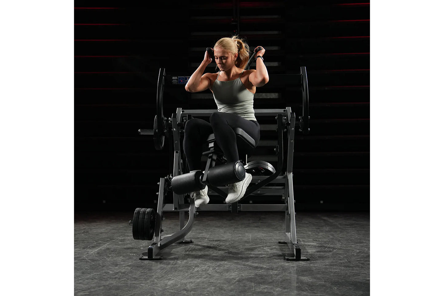 Woman in gray tank top and black leggings performing abdominal exercise on Hammer Strength plate-loaded machine in gym settin