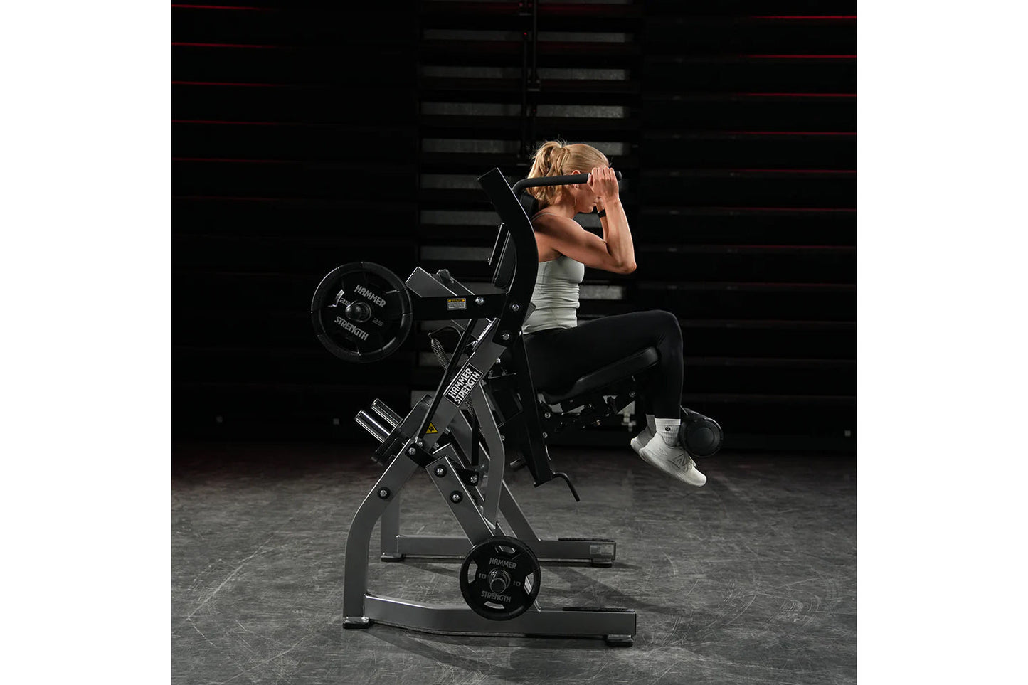 Woman using black Hammer Strength plate-loaded abdominal oblique crunch machine in gym setting, wearing white tank top and sn