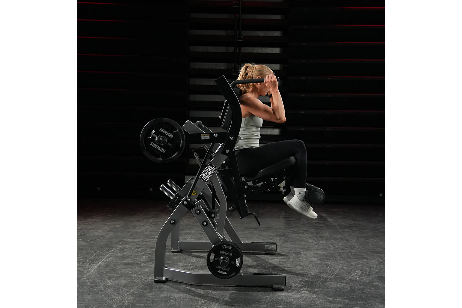 Woman using black Hammer Strength plate-loaded abdominal oblique crunch machine in gym setting, wearing white tank top and sn