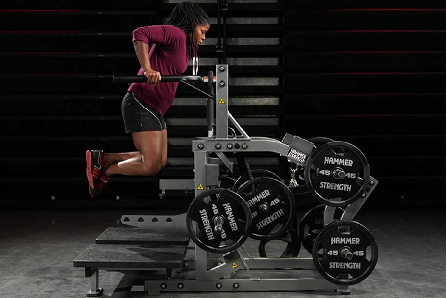 Woman in burgundy t-shirt and black shorts using Hammer Strength plate-loaded belt squat machine with weight plates, stairs i