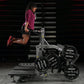 Woman using burgundy long-sleeve shirt and black shorts on plate-loaded belt squat machine with weight plates, indoor gym set