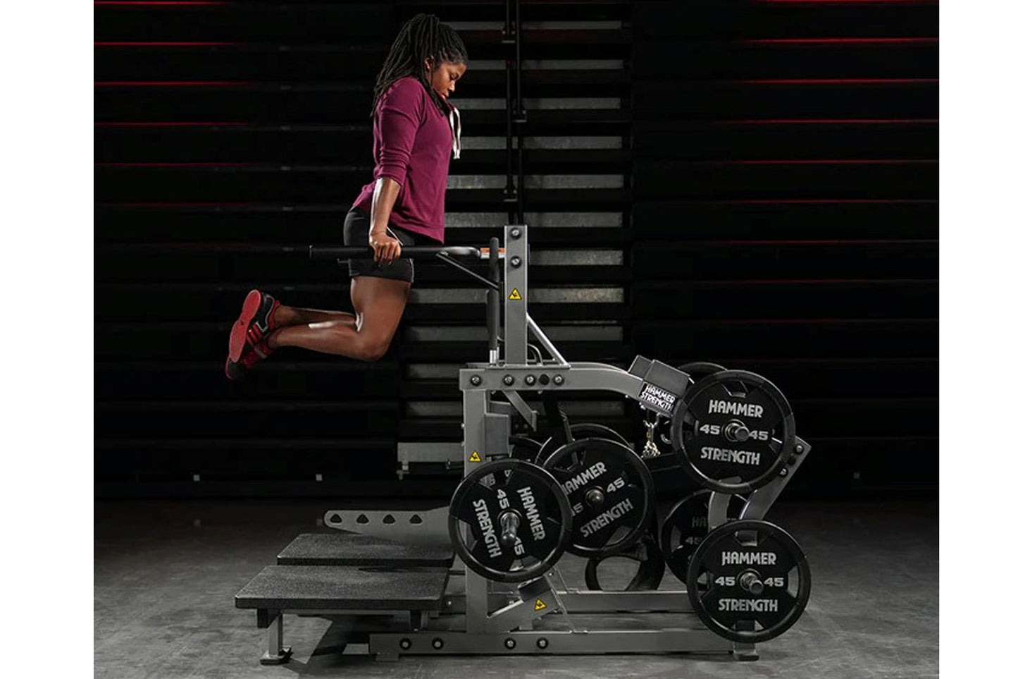 Woman using burgundy long-sleeve shirt and black shorts on plate-loaded belt squat machine with weight plates, indoor gym set