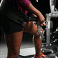 Person in burgundy tank top and black shorts using plate-loaded belt squat machine with silver chain attachment in gym settin