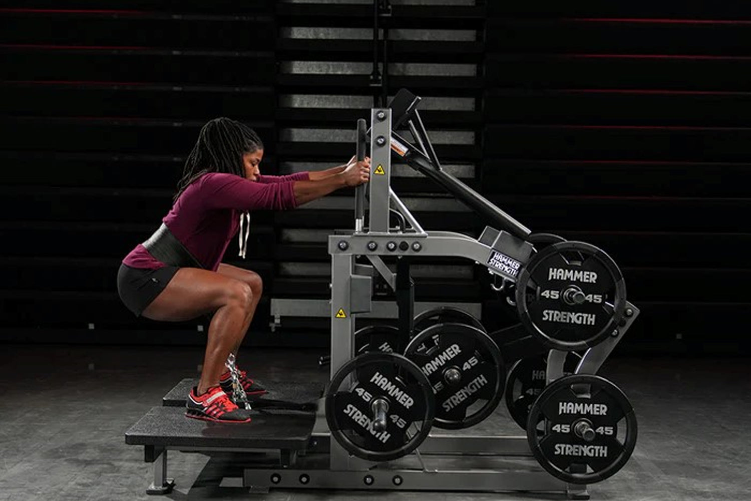 Woman in burgundy top performing leg exercise on Hammer Strength plate-loaded belt squat machine with weight plates in gym se