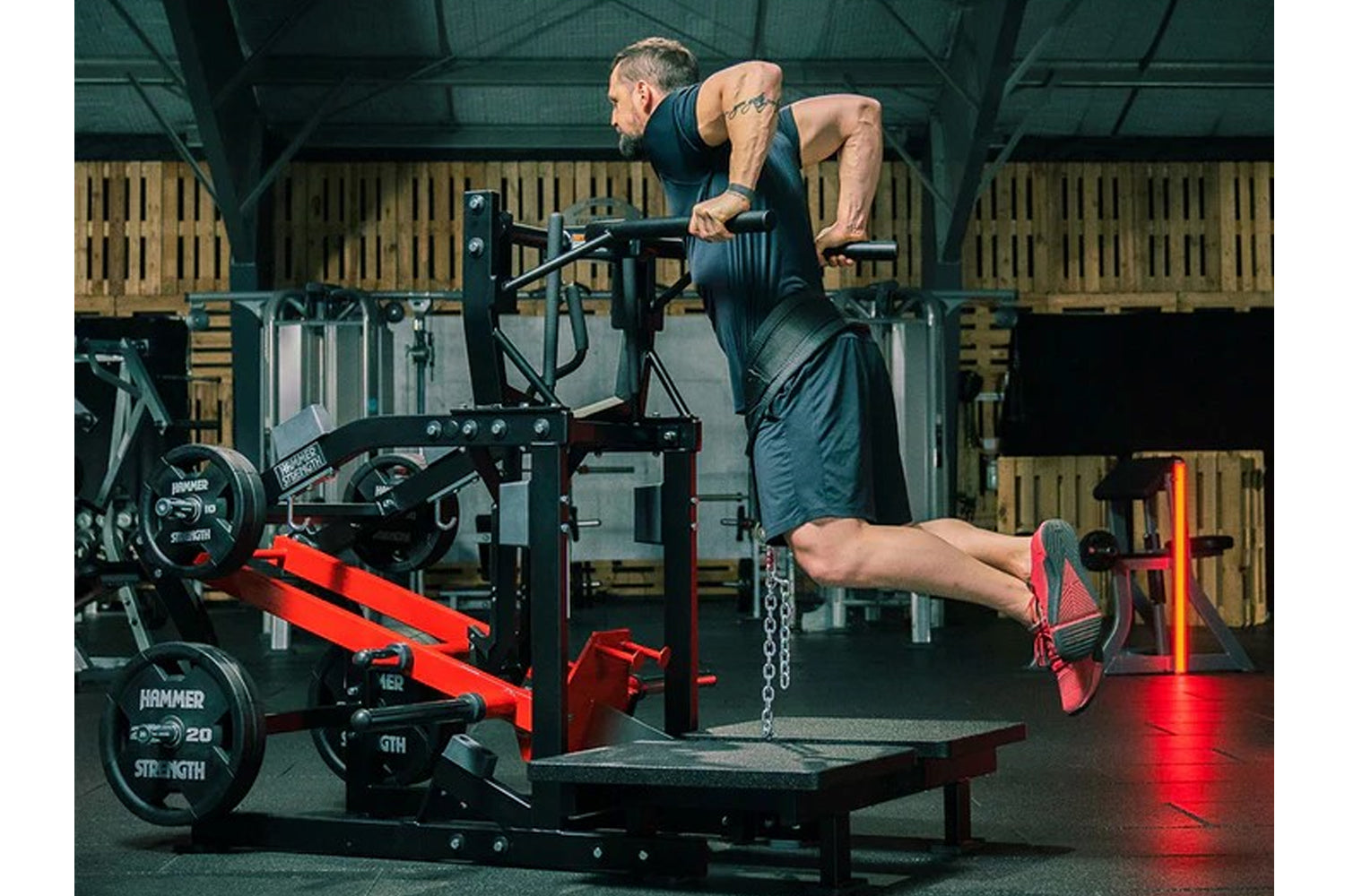Man using Hammer Strength plate-loaded belt squat machine in gym, wearing dark tank top and red sneakers