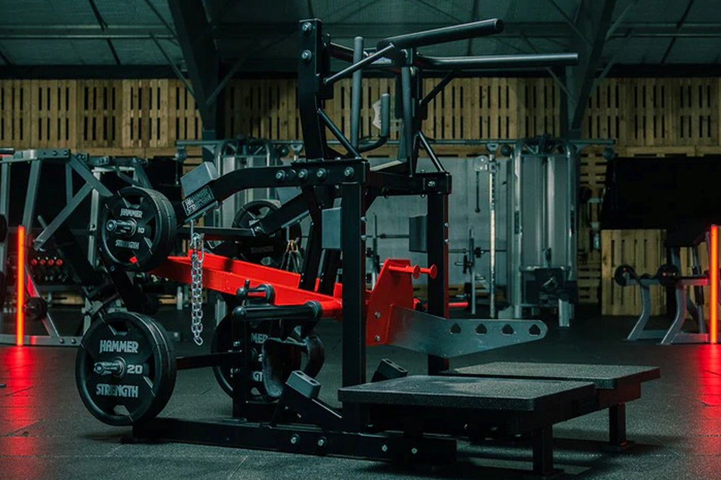 Black and red plate-loaded belt squat machine with chain attachment and foot platform in gym setting