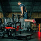 Man in dark blue athletic shirt and shorts using red and black plate-loaded belt squat machine in gym setting