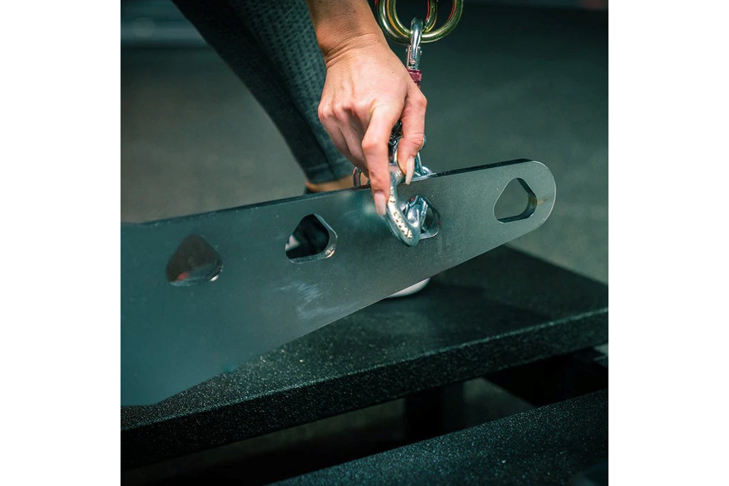 Hand attaching carabiner clip to dark green metal belt squat machine attachment plate with multiple anchor holes