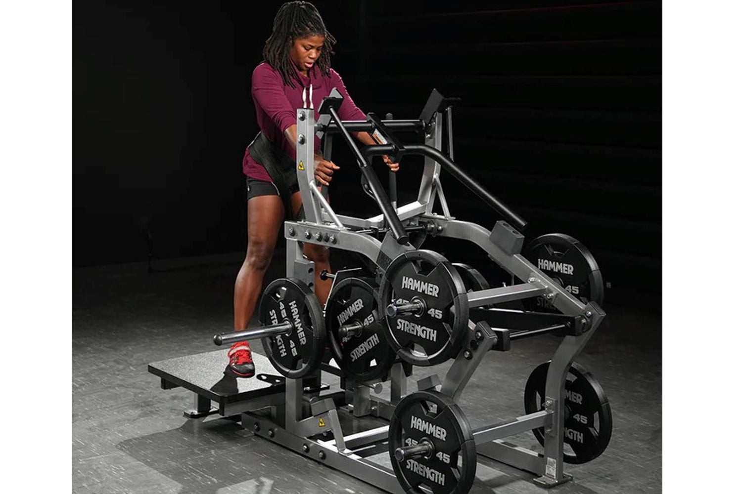Woman in burgundy hoodie and black shorts using silver and black Hammer Strength plate-loaded belt squat machine in gym setti