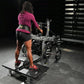 Woman wearing magenta long-sleeve top and black shorts using plate-loaded belt squat machine in gym setting