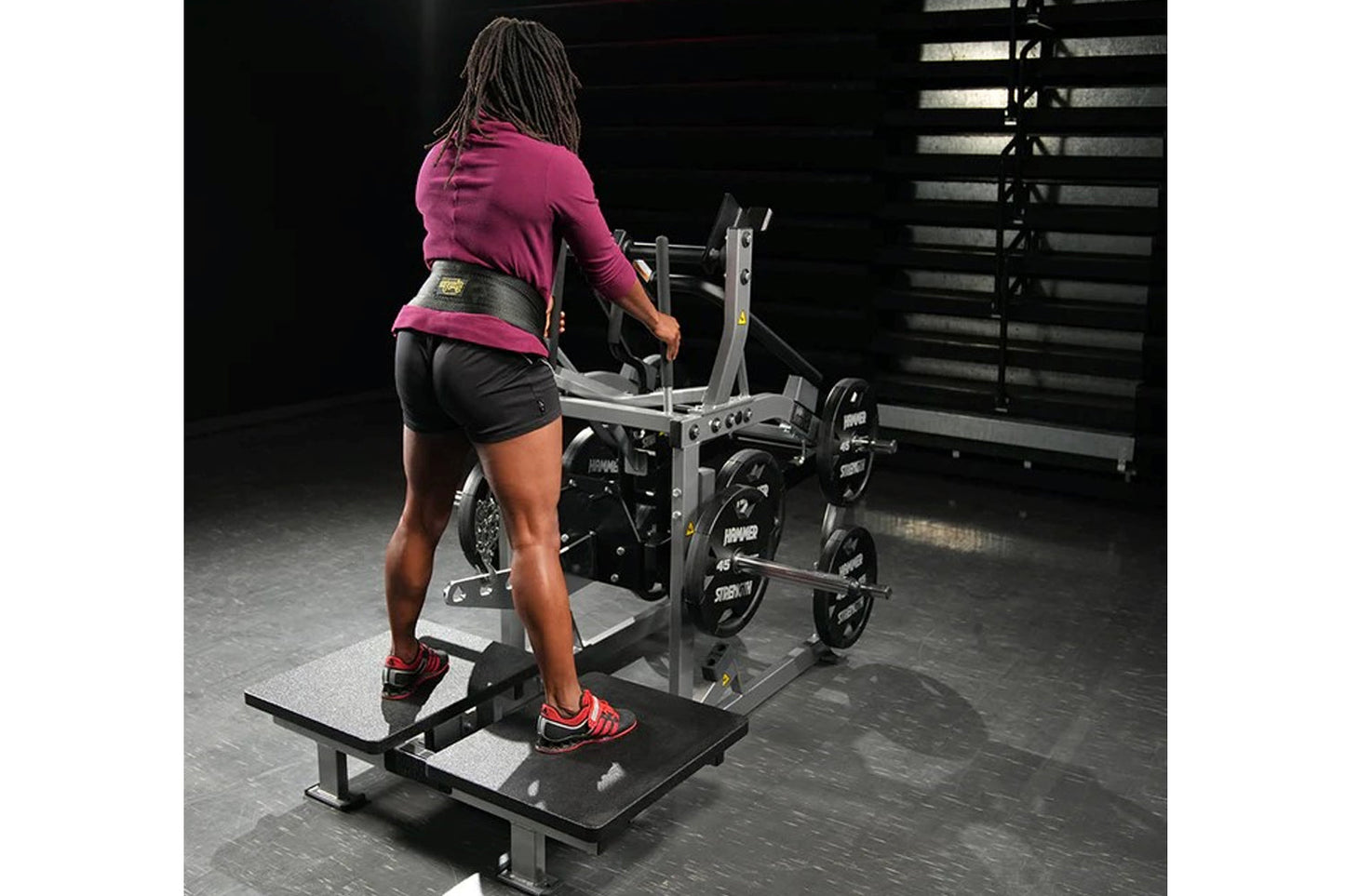 Woman wearing magenta long-sleeve top and black shorts using plate-loaded belt squat machine in gym setting