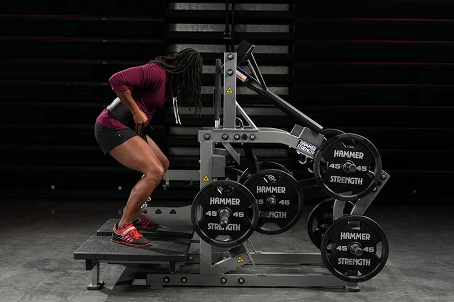Athlete using Hammer Strength plate-loaded belt squat machine with 45-pound weight plates, wearing burgundy top and red sneak