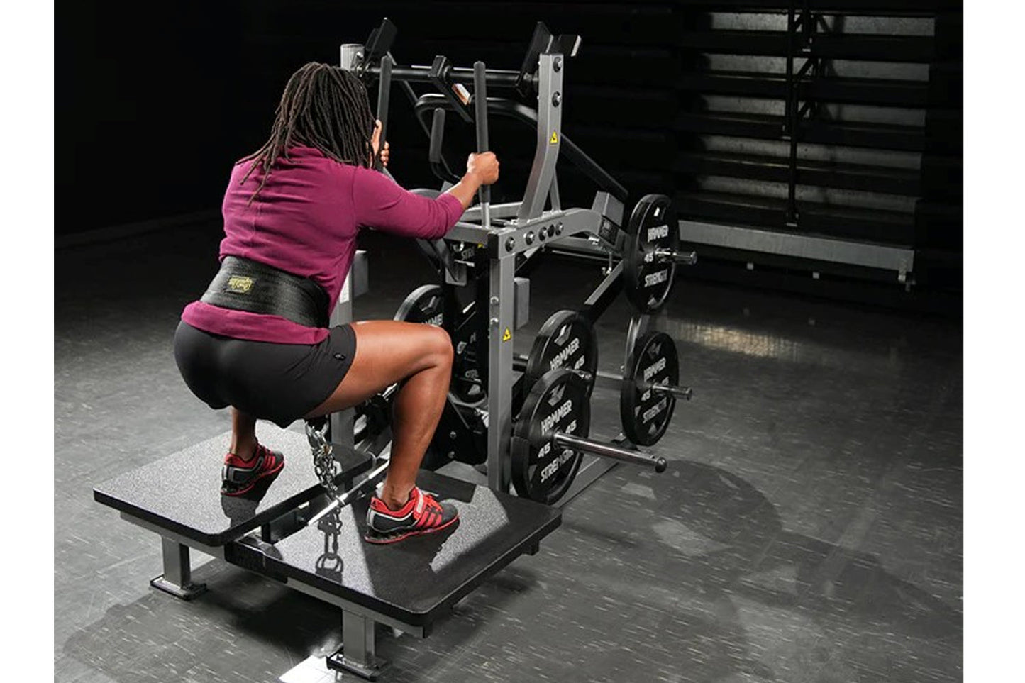 Woman using black Hammer Strength plate-loaded belt squat machine in gym setting, wearing magenta long-sleeve top and black s
