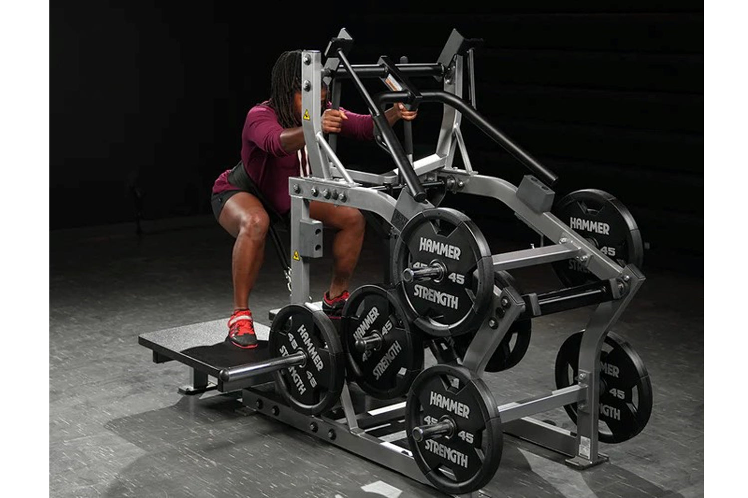 Person in burgundy top using Hammer Strength plate-loaded belt squat machine with black weight plates, on gray floor