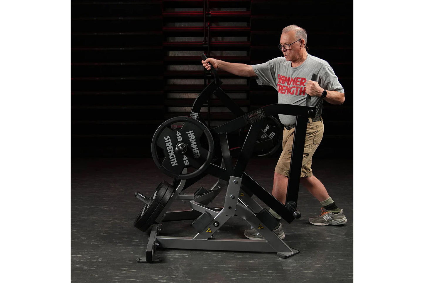Man wearing gray Hammer Strength t-shirt using black plate-loaded exercise machine with weighted wheel in gym setting