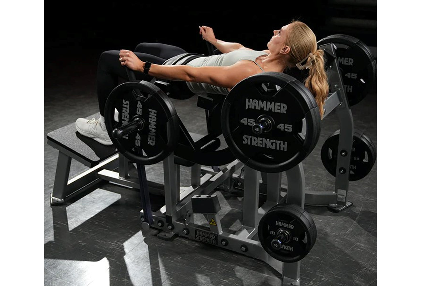 Woman using black Hammer Strength plate-loaded glute drive machine with 45-pound plates in gym setting