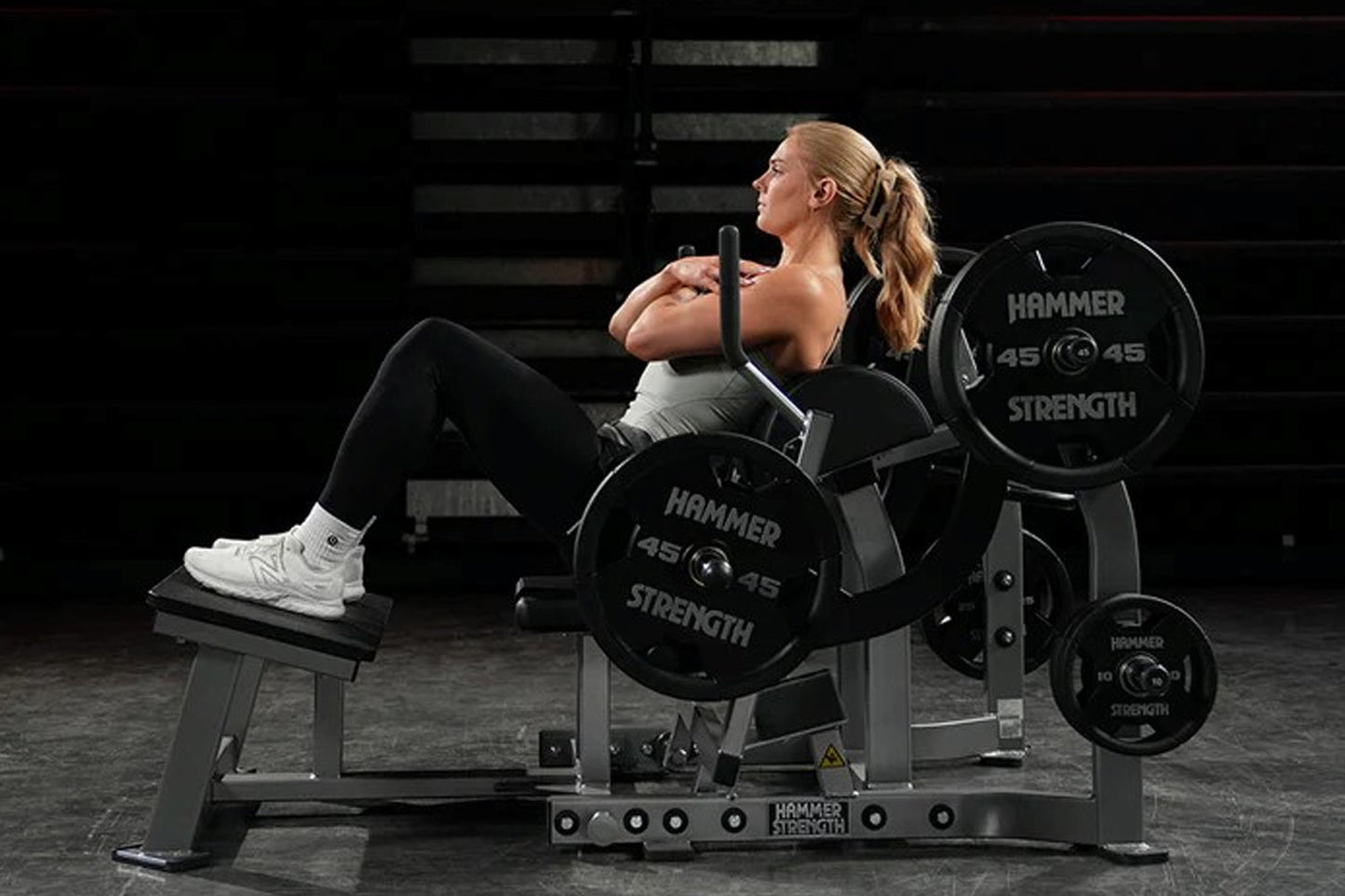 Woman using black Hammer Strength plate-loaded glute drive machine with loaded weight plates in gym setting