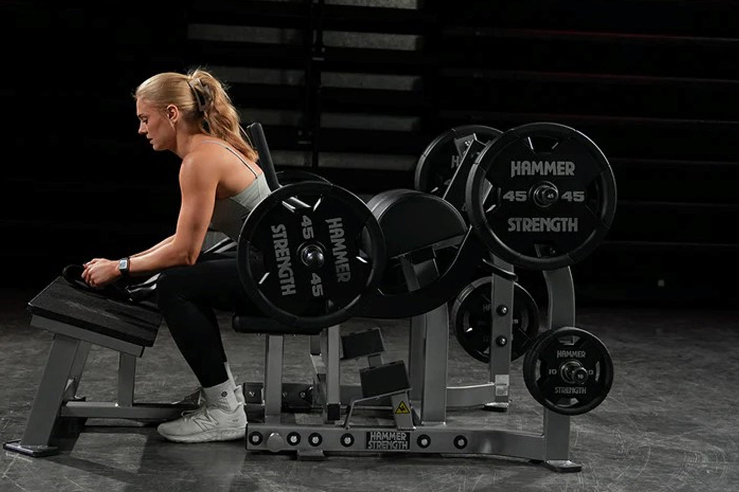 Blonde woman in gray sports bra and black pants performing glute drive exercise on Hammer Strength black plate-loaded machine