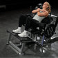 Woman using black and gray plate-loaded glute drive machine in gym setting, wearing gray tank top and black leggings