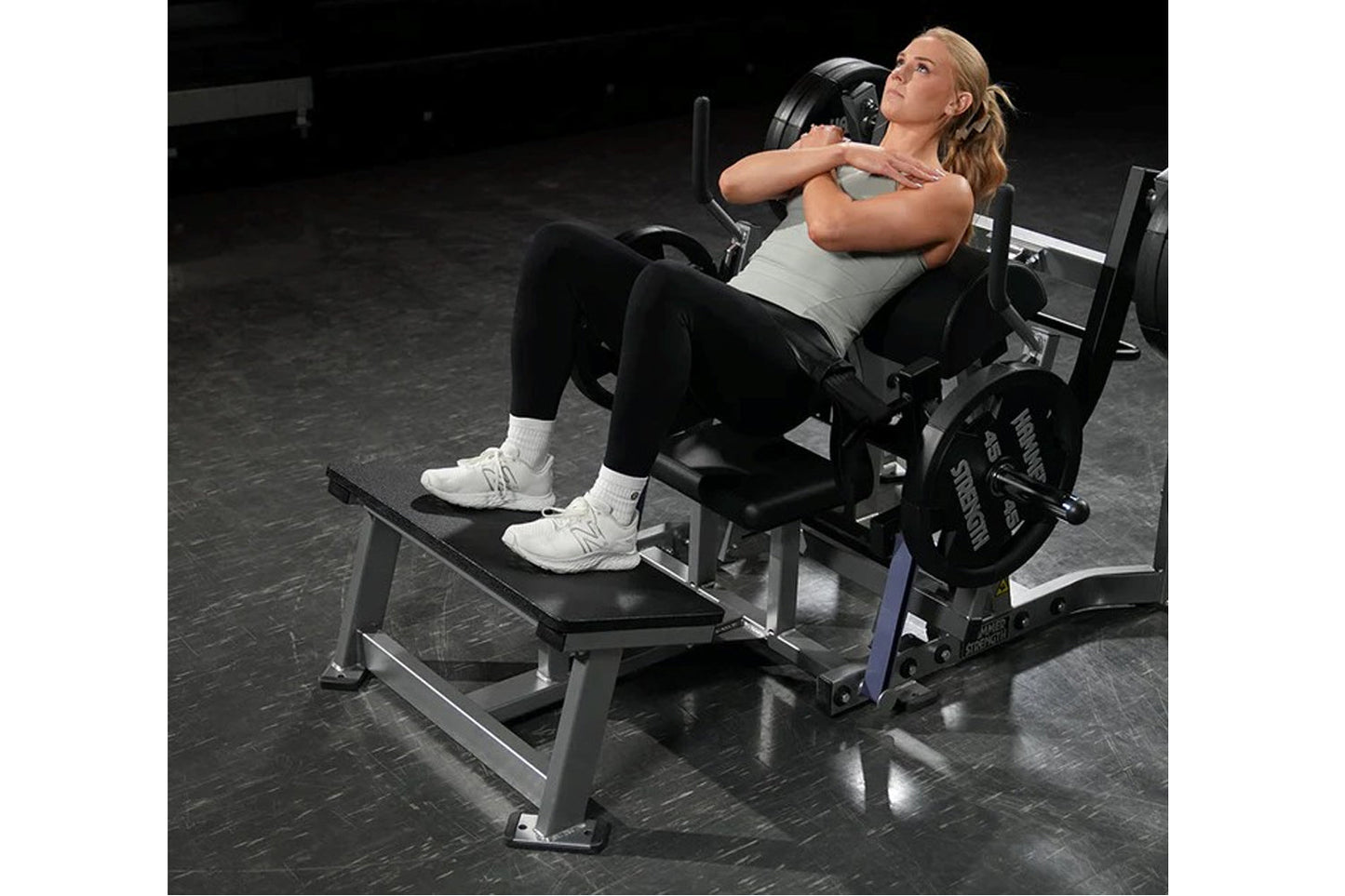 Woman using black and gray plate-loaded glute drive machine in gym setting, wearing gray tank top and black leggings