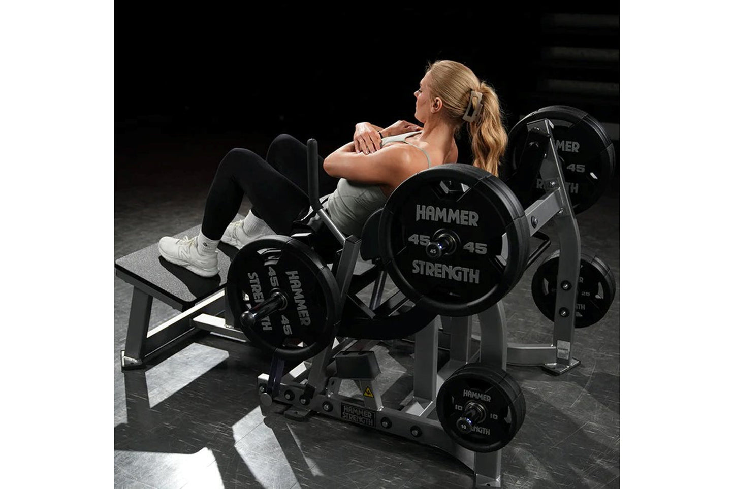 Woman using Hammer Strength plate-loaded glute drive machine with black weight plates in gym setting