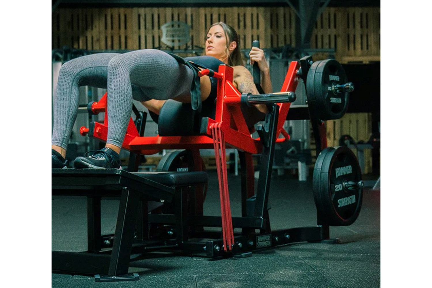 Woman in gray textured leggings using red Hammer Strength plate-loaded glute drive machine in gym setting