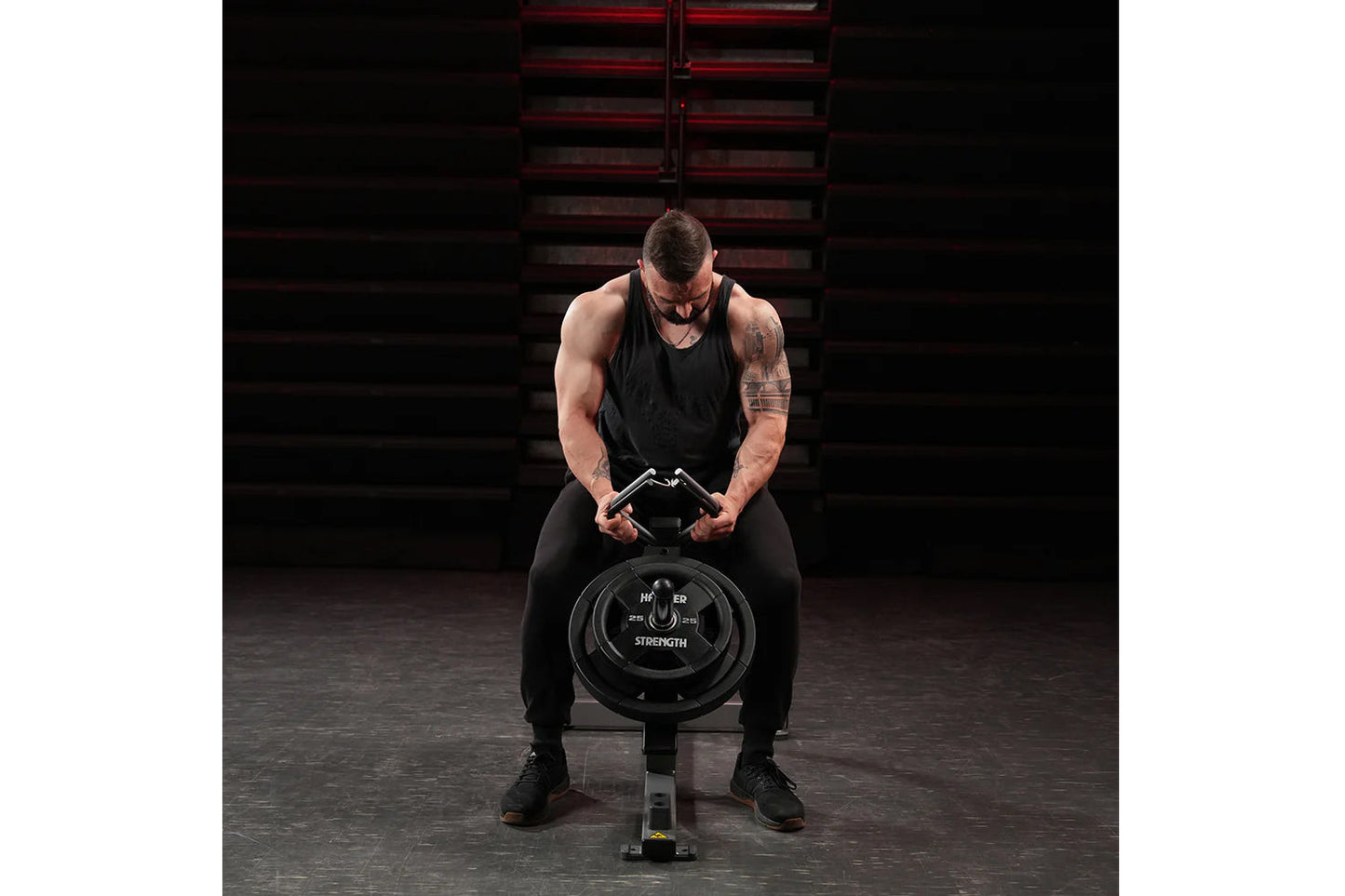 Man in black tank top and pants using Hammer Strength plate-loaded gripper in gym setting with red metal stairs background