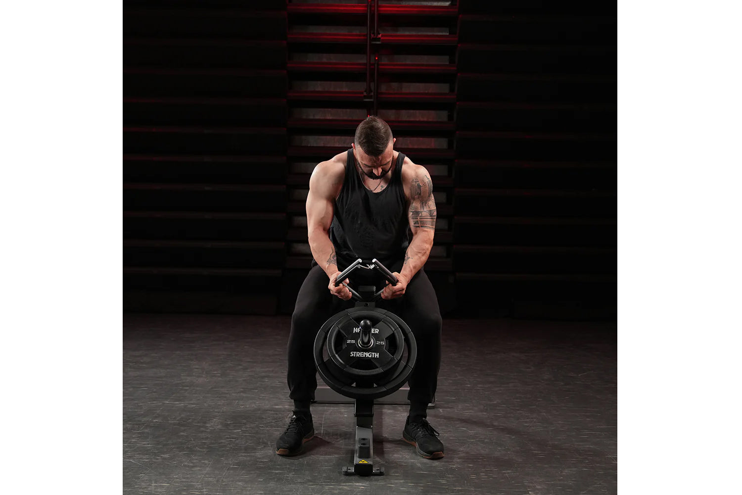 Man in black tank top and pants using Hammer Strength plate-loaded gripper in gym setting with red metal stairs background