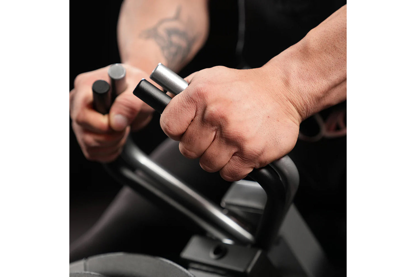 Person gripping black and silver plate-loaded gripper with cylindrical handles, demonstrating exercise equipment on dark back