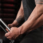 Person with tattooed arms wearing black long-sleeve shirt, holding metal plate-loaded gripper exercise equipment