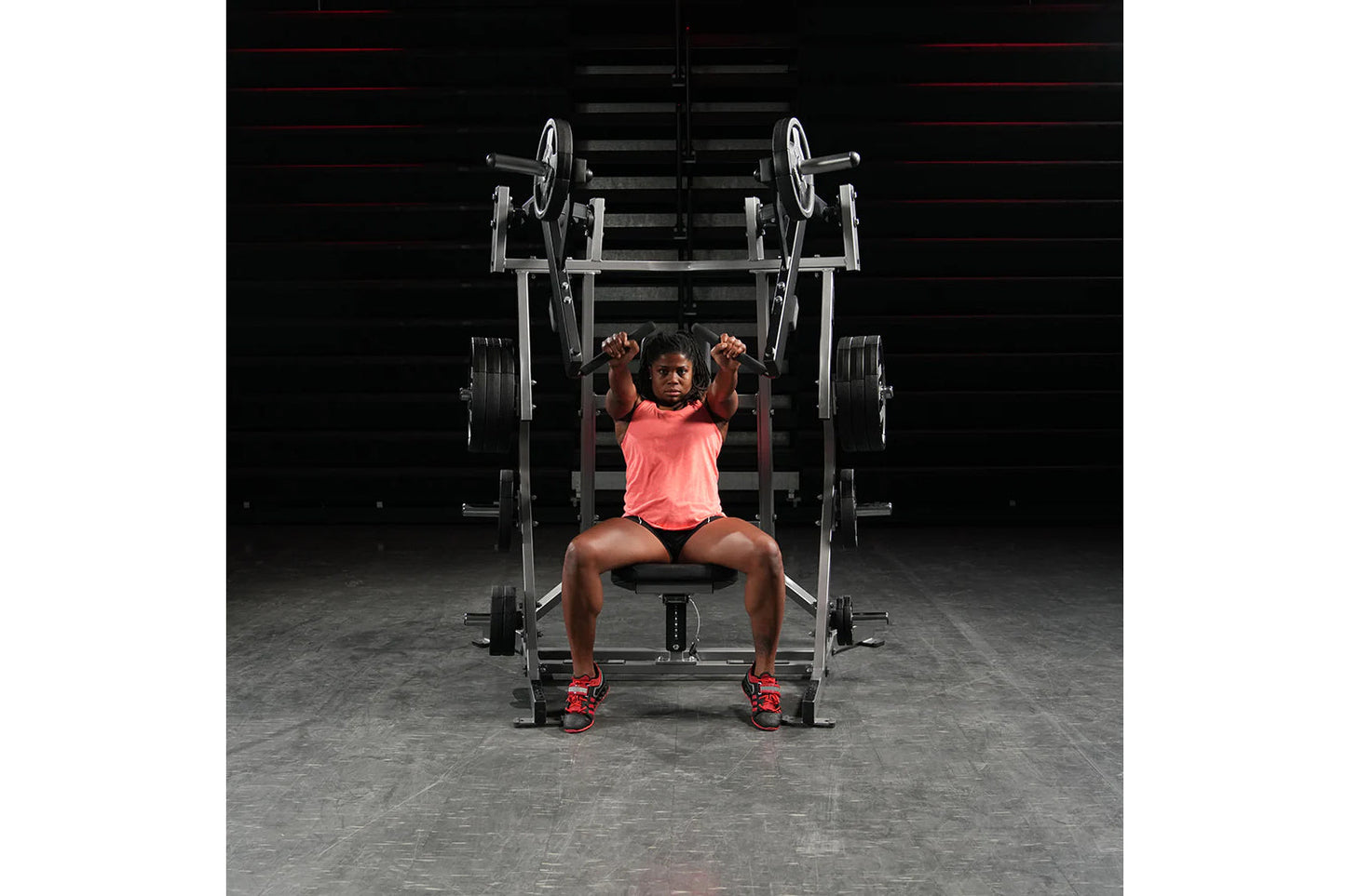 Woman in coral tank top and black shorts performing chest press on silver and black plate-loaded bench press machine in gym
