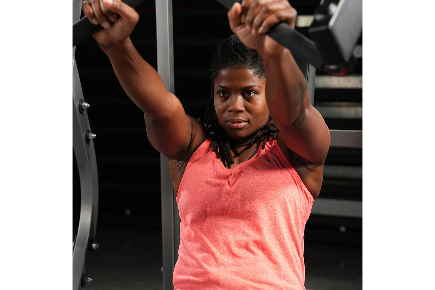Woman wearing coral sleeveless tank top using plate-loaded bench press machine in gym setting