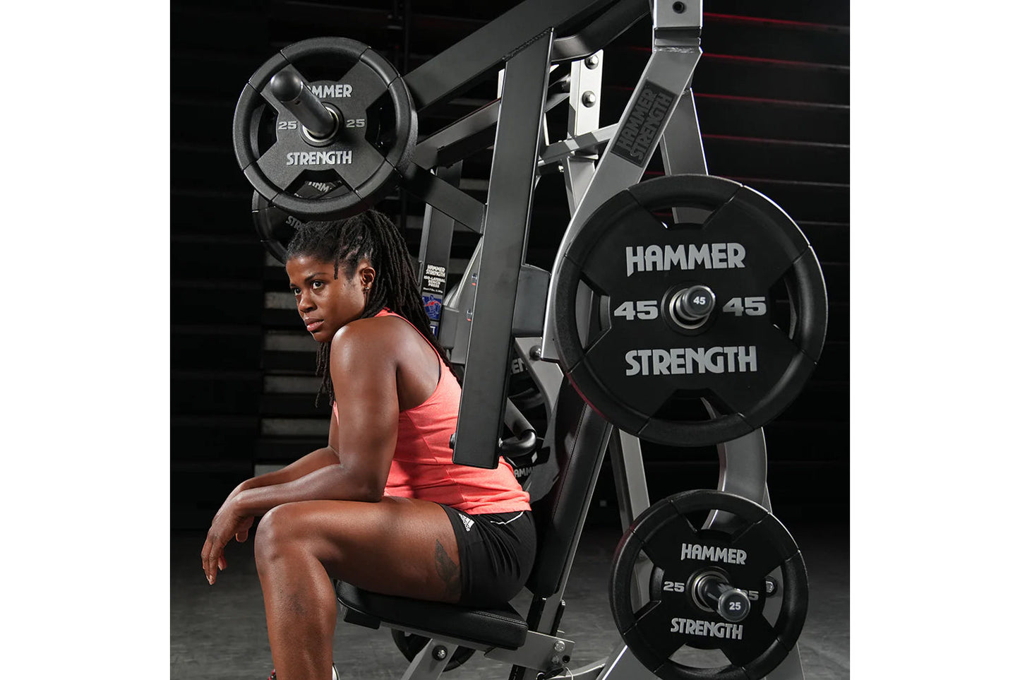 Woman in coral tank top and black shorts seated on Hammer Strength plate-loaded bench press machine with black weight plates