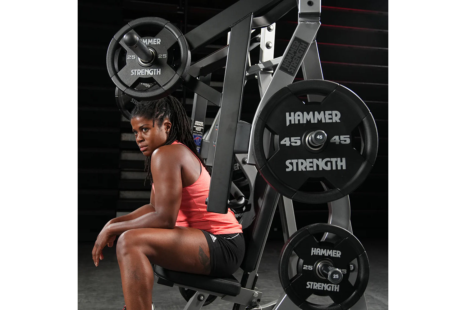 Woman in coral tank top and black shorts seated on Hammer Strength plate-loaded bench press machine with black weight plates