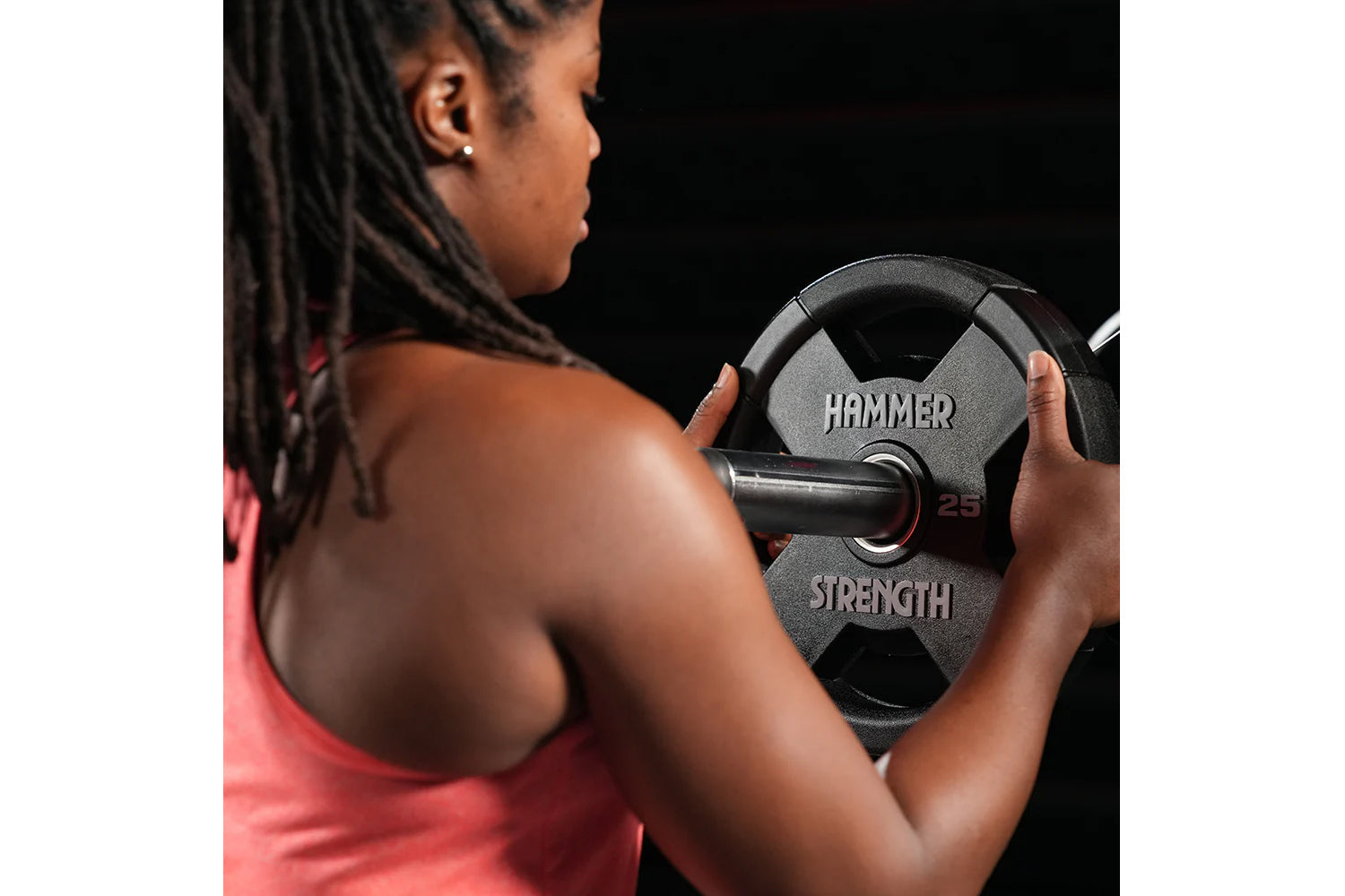 Woman in pink tank top holding black and gray Hammer Strength 25-pound dumbbell against black background