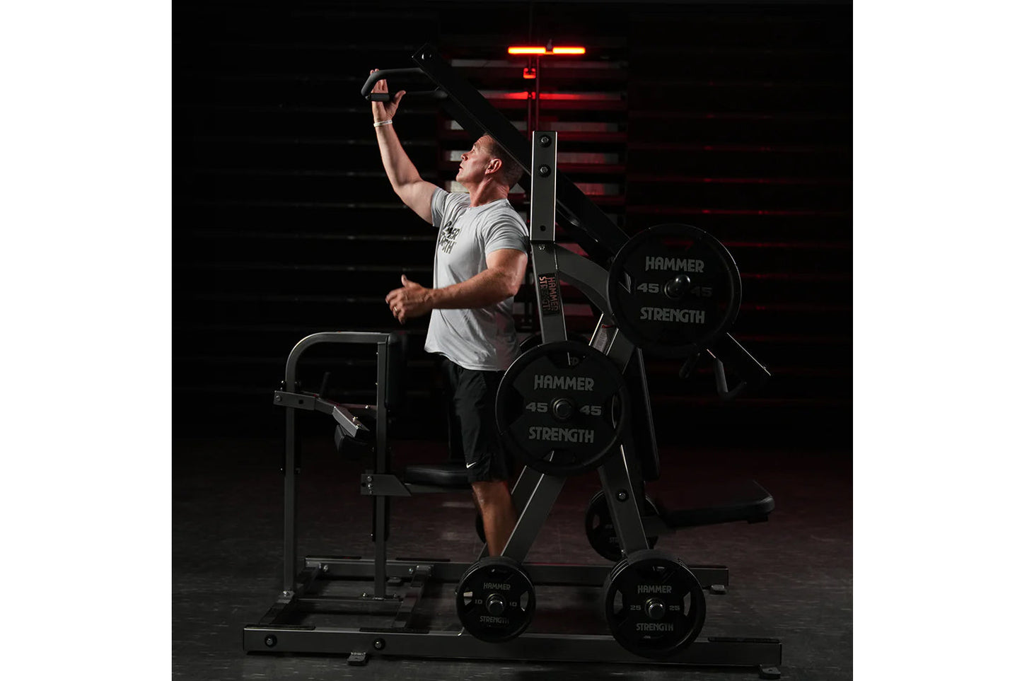Man in light gray t-shirt using black Hammer Strength plate-loaded chest/back machine with red neon lighting in dark gym sett