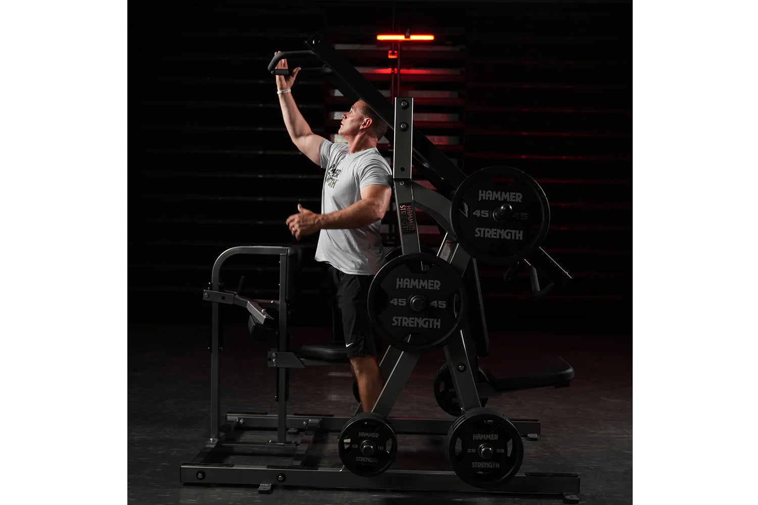 Man in light gray t-shirt using black Hammer Strength plate-loaded chest/back machine with red neon lighting in dark gym sett