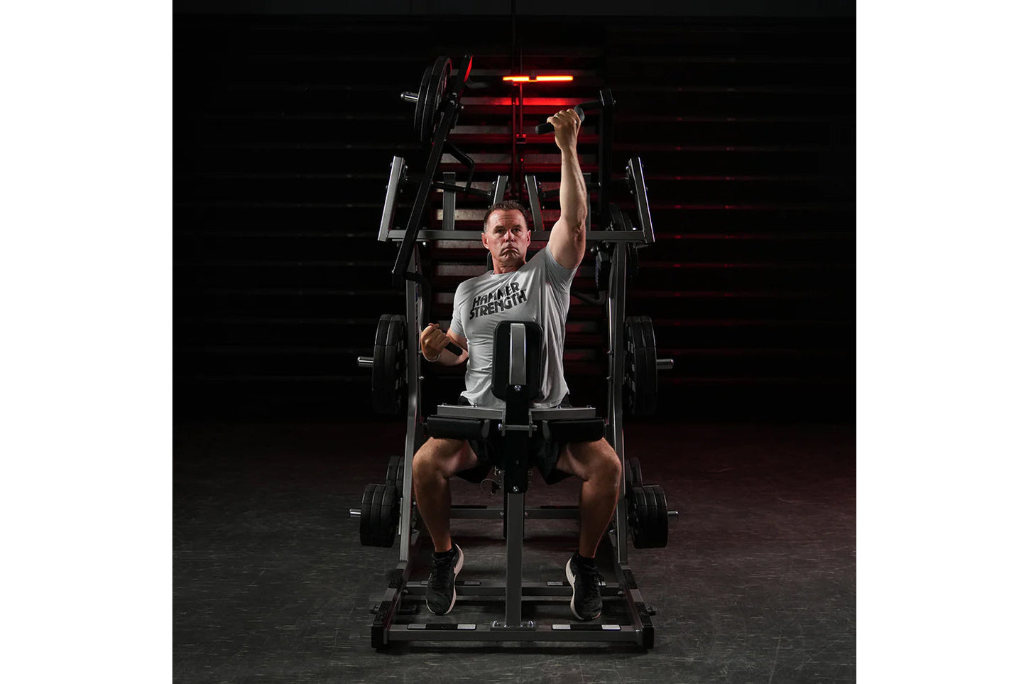 Man wearing gray "Hammer Strength" t-shirt performing chest press exercise on black plate-loaded machine with red neon lights